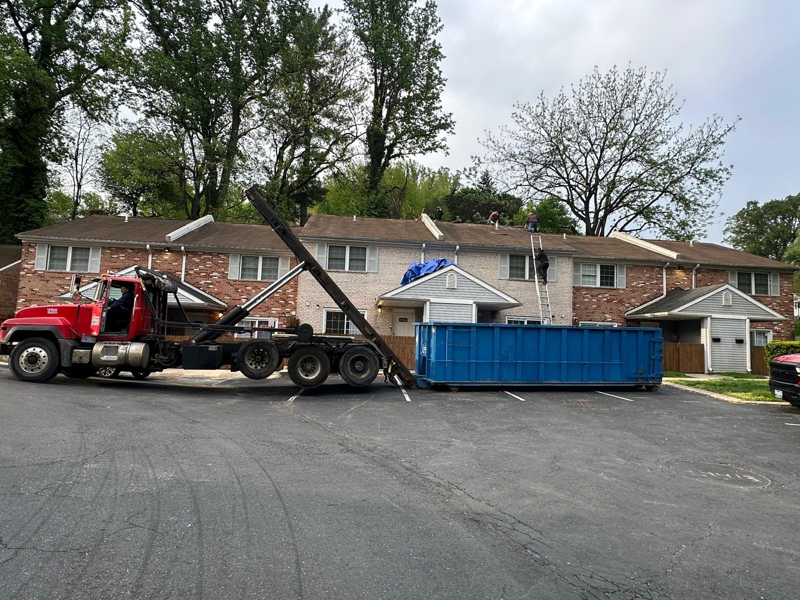 Workers on the roof of a residential building refurbishing or repairing the roof, with a crane and dumpster in the parking lot in front.