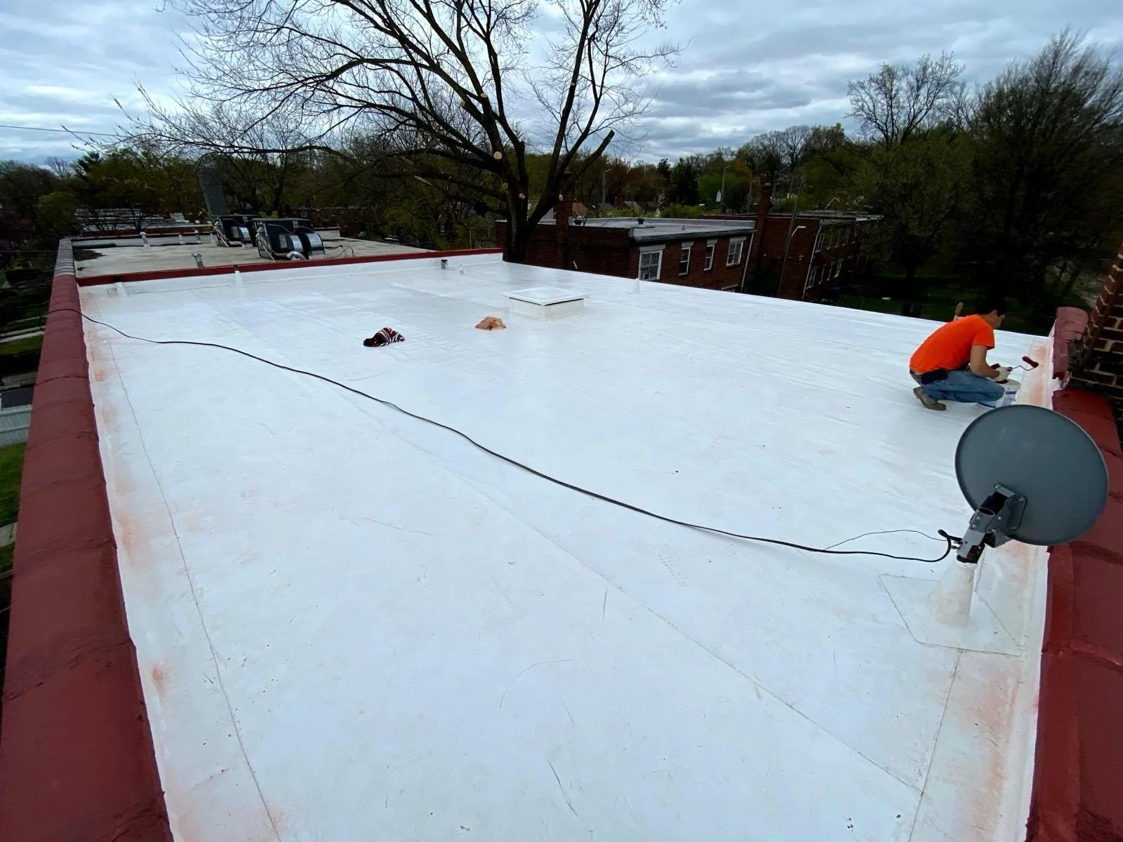 A person working on a flat rooftop, which has been painted white, with a tree and neighboring rooftops in the background.