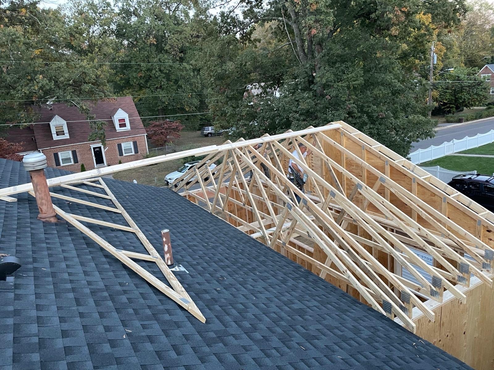 View from a rooftop showing a house under construction, with wooden framing for a new roof. A crew of workers is on the framing, and the background includes a large tree, neighboring houses, and a street with parked cars.