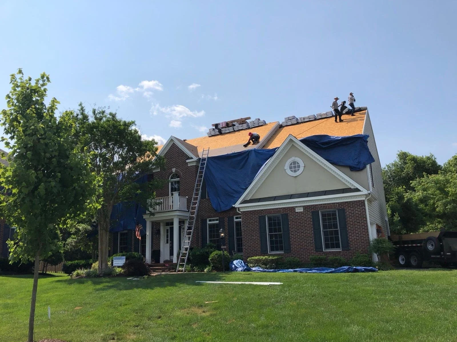 Workers installing a new roof on a brick house with a front porch, surrounded by greenery, on a clear day.
