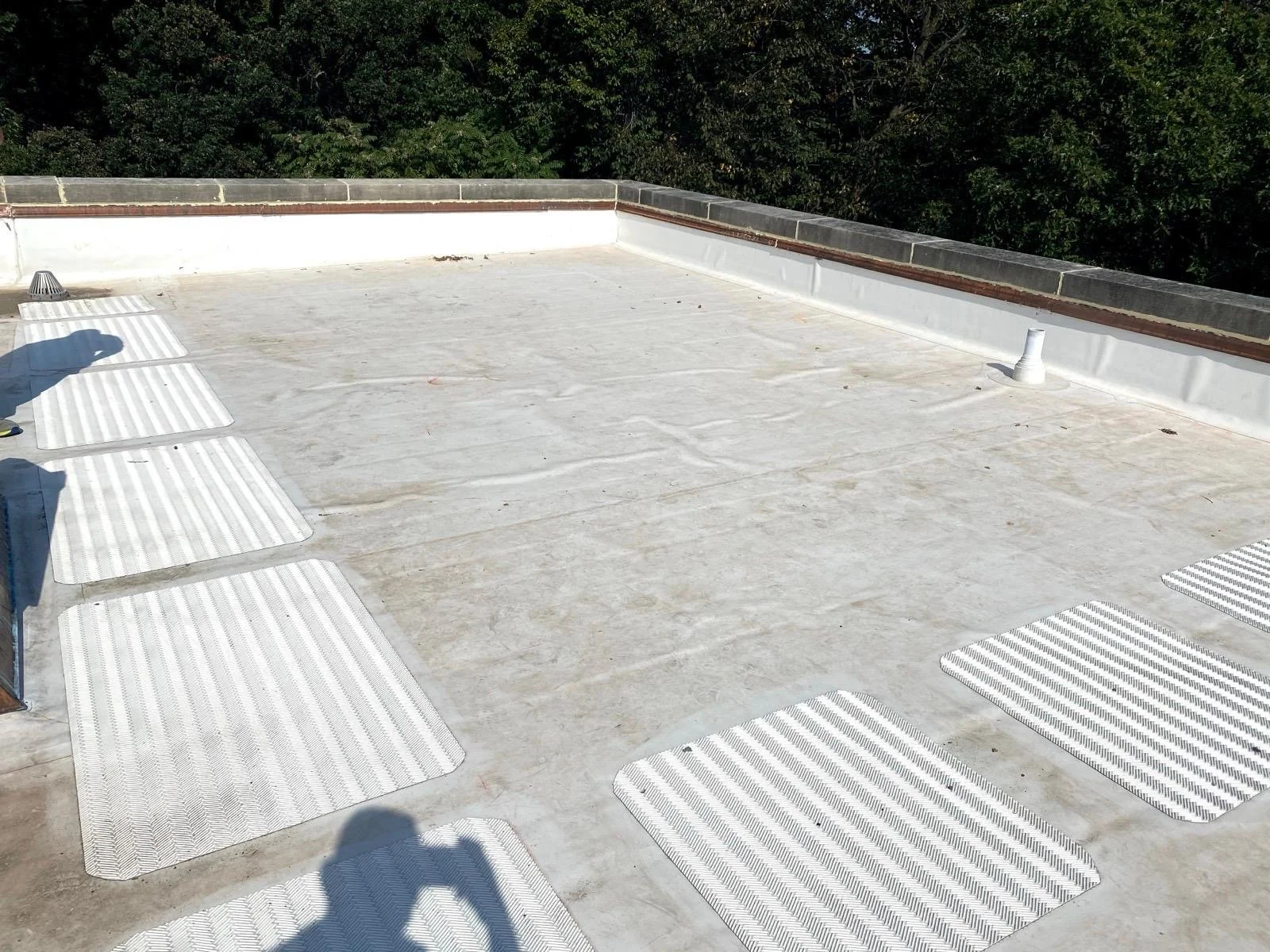 Flat rooftop with several white and gray striped mats and a person's shadow in the lower left corner. The background shows trees and a brick and gray stone wall border.
