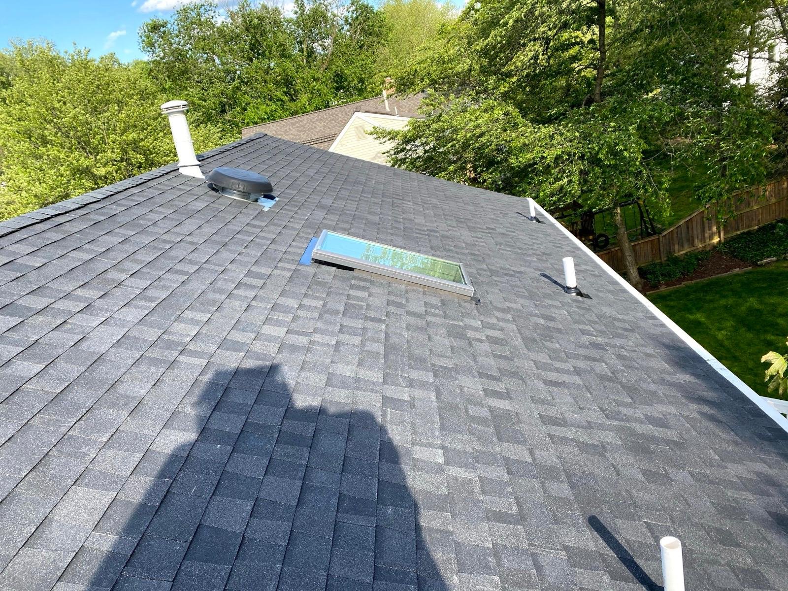 View of a house roof with dark gray shingles, a skylight window, a vent, and several pipes. Green trees and neighboring houses are visible in the background on a sunny day.