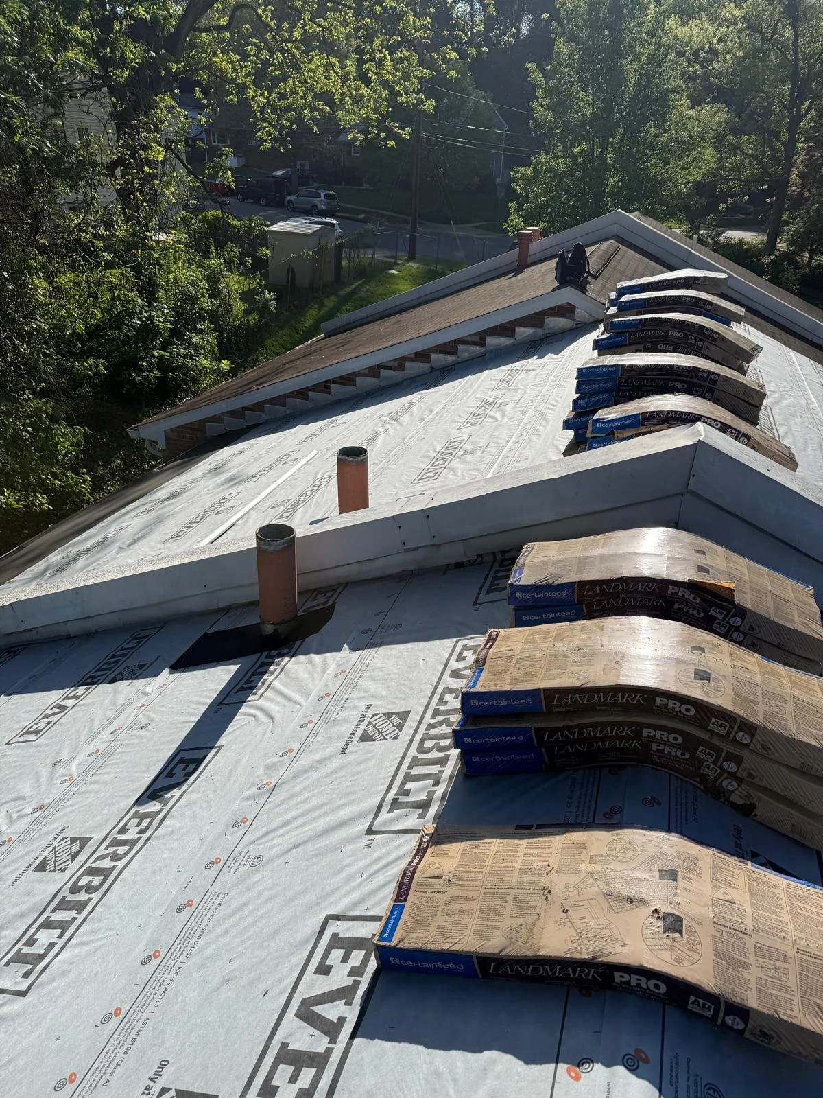 Roof under construction with insulation material and roofing shingles stacked on the ridge. The roof has two sections, one partially finished with shingles and the other covered with white underlayment. Chimney pipes protrude from the roof, and a pro