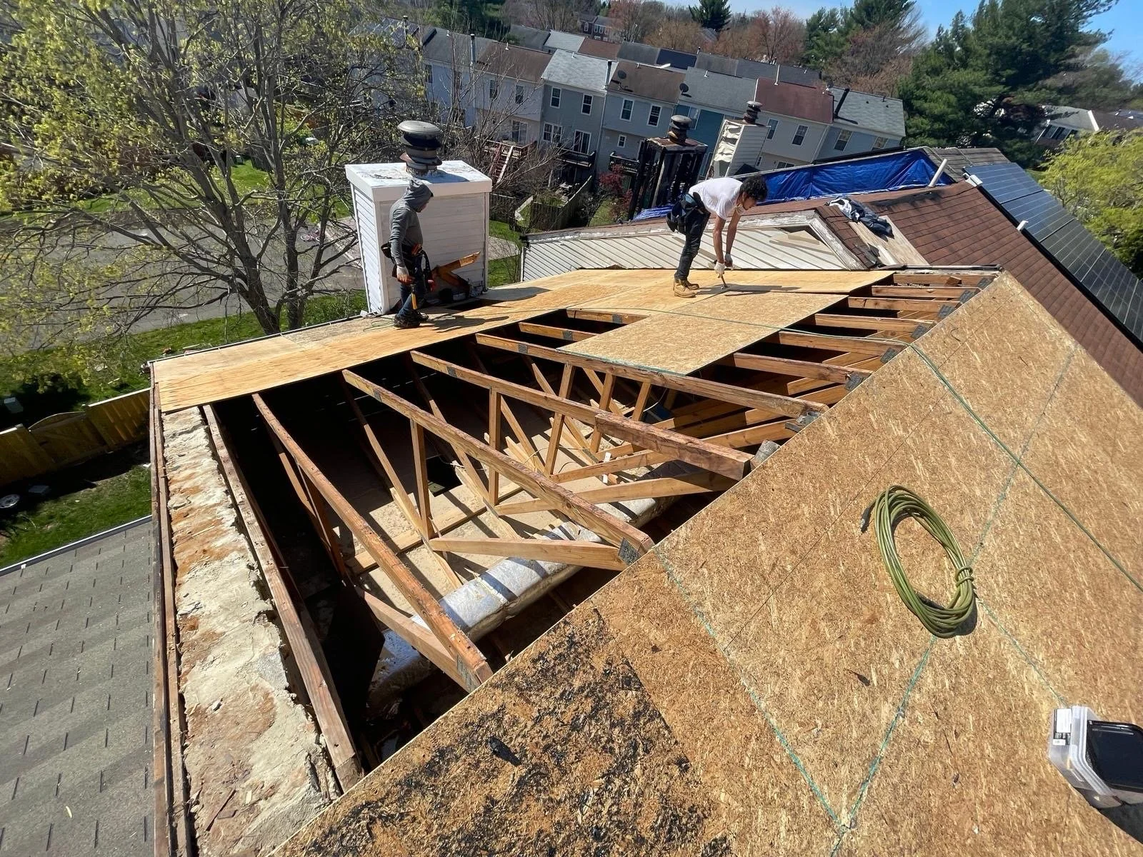 Two workers are on the roof of a house under construction, installing new wooden sheathing and rafters. One worker is standing and using a tool, while the other is bent over, working on the roof. The roof has some completed sections with shingles, an