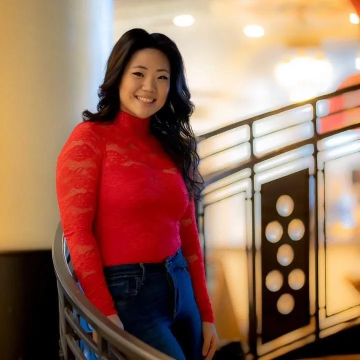 A woman smiling, wearing a red lace long-sleeve top and blue jeans, standing near a decorative railing in an indoor setting with warm lighting.