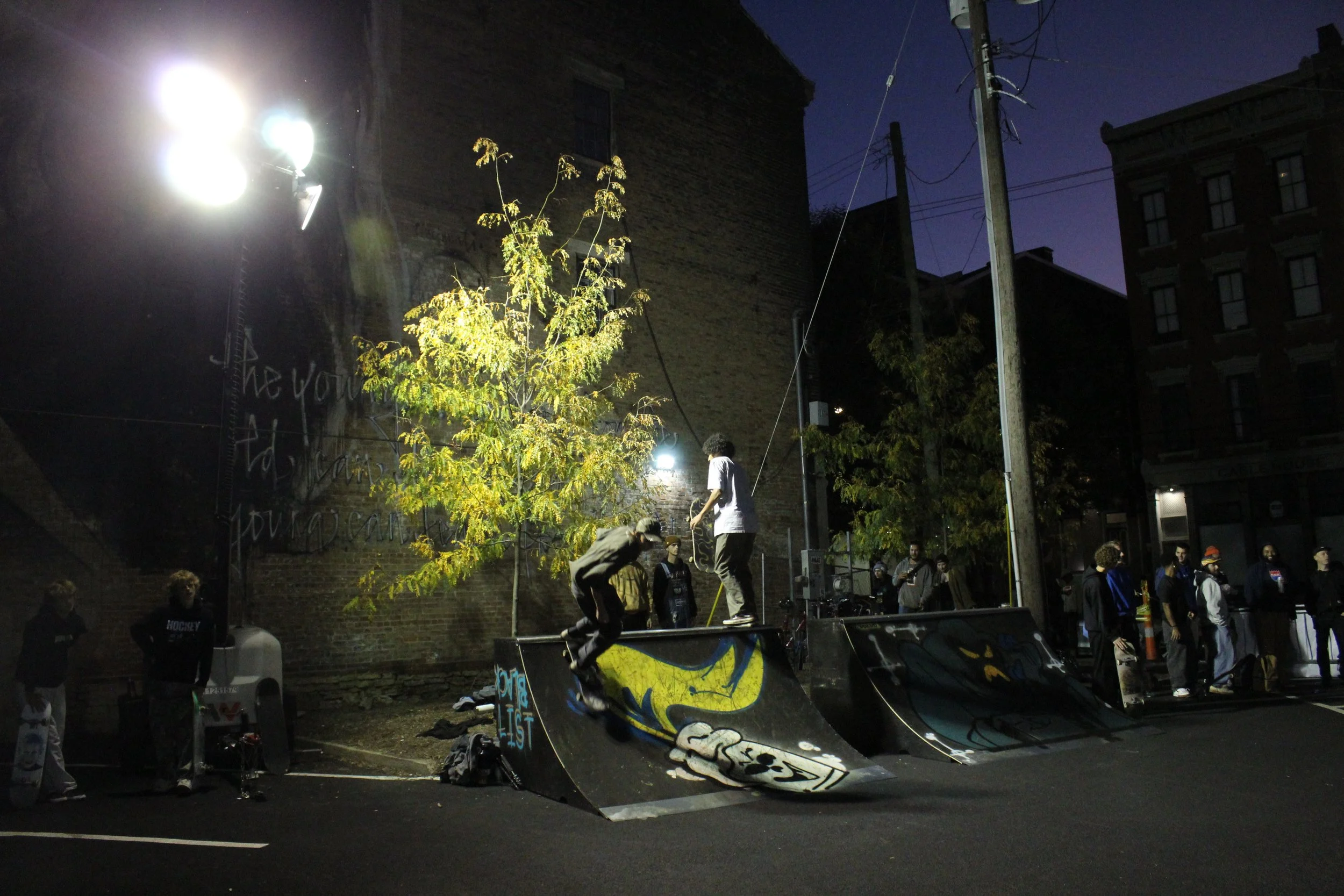 People skateboarding at an outdoor skate park at night with a brick building, utility poles, and a small tree illuminated by artificial lights in the background.