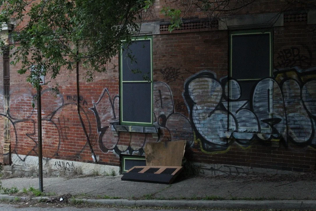 Urban alleyway with graffiti-covered brick wall, a small tree, two windows, and a wooden pallet leaning against the wall.