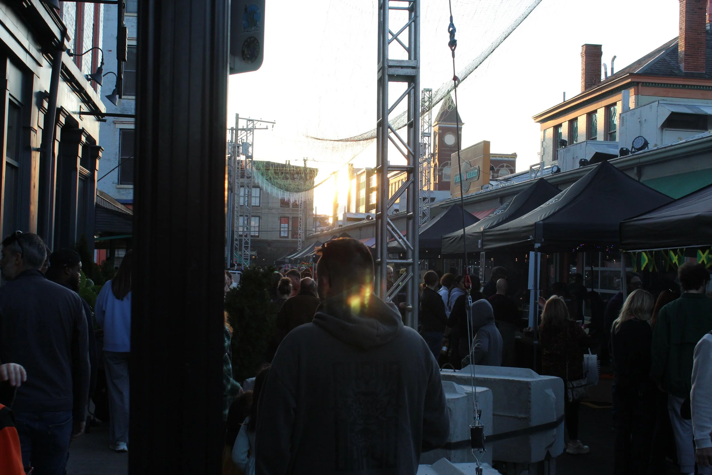 An outdoor urban market scene during sunset with a crowd of people standing and walking under tents, buildings, and a clock tower in the background.
