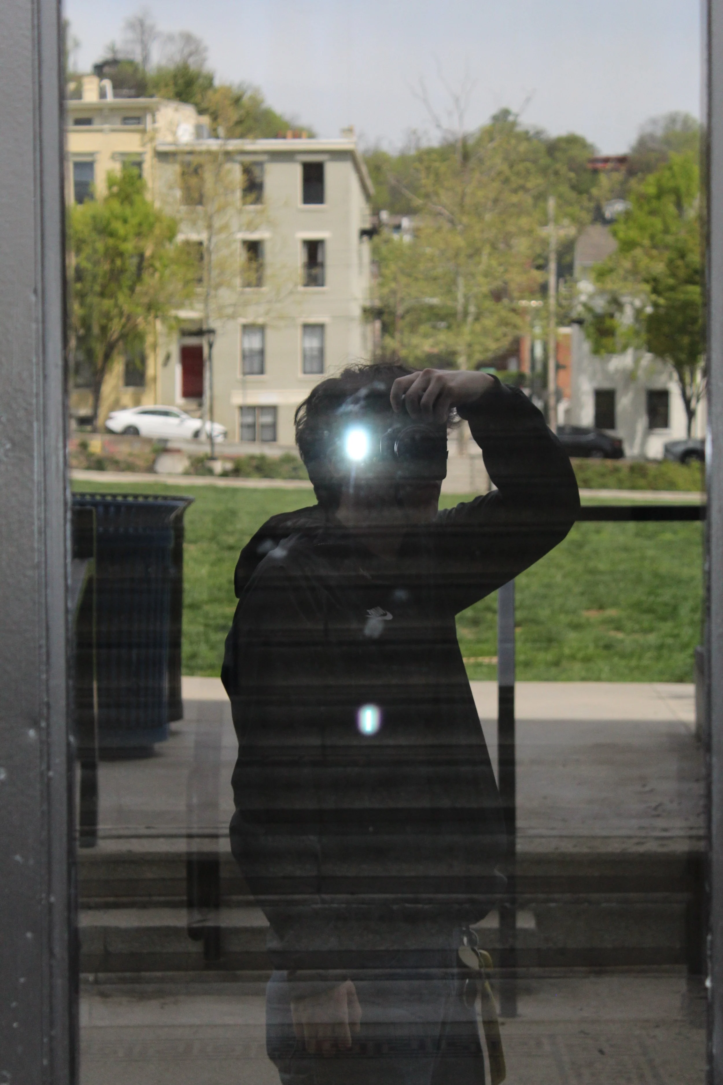 Reflection of a person taking a photo with a camera or phone in a window or glass door, with a background of trees, buildings, and cars outside.