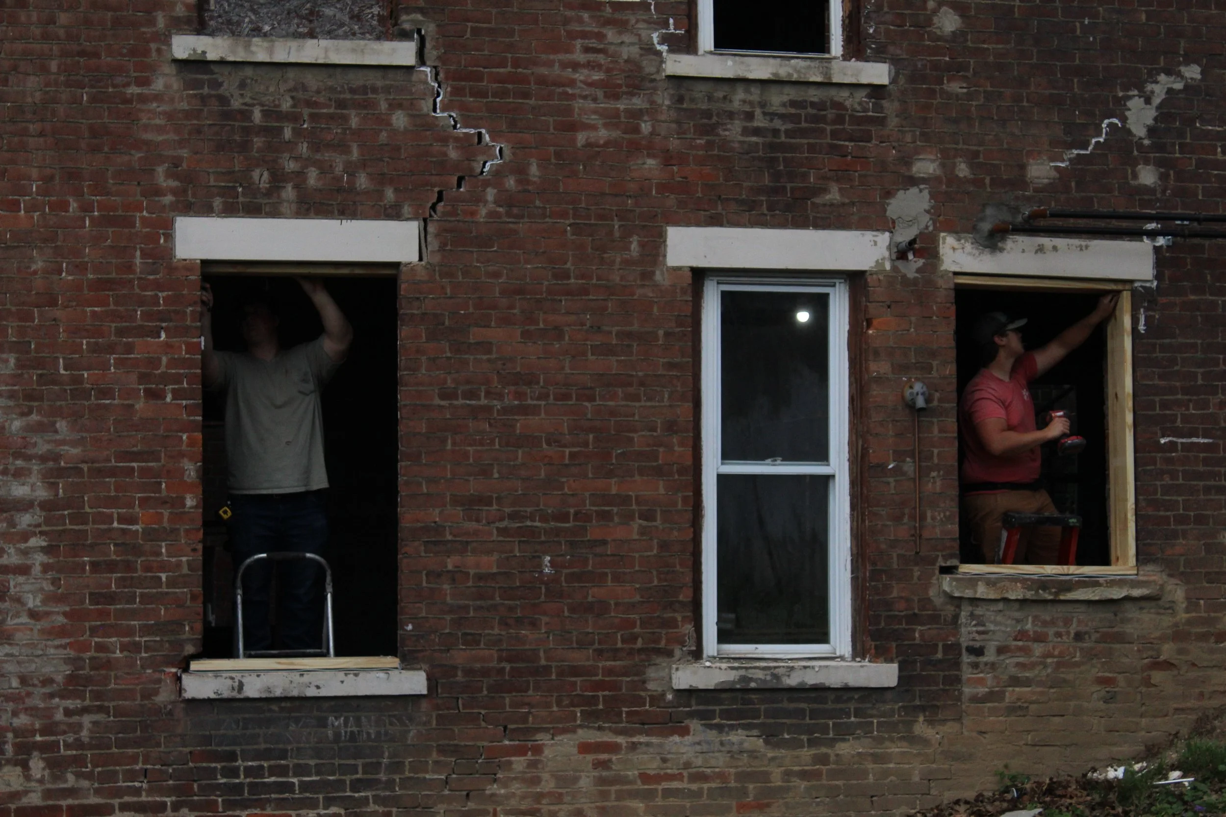 Two men doing construction work on a brick building, one inside a window opening and the other at a doorway, with tools and construction materials around.