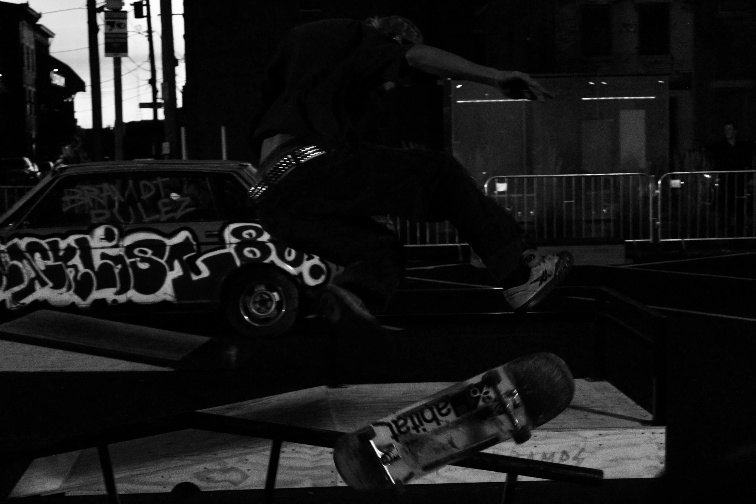 Black and white photo of a person wearing a hoodie riding a skateboard on a city street at night, with a graffiti-covered car and construction barriers in the foreground and background.