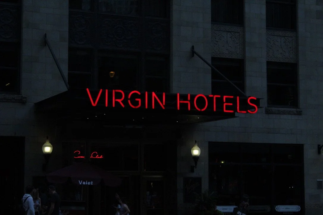 The entrance of Virgin Hotels with a neon sign reading "VIRGIN HOTELS" above the doorway, and people walking outside at dusk.