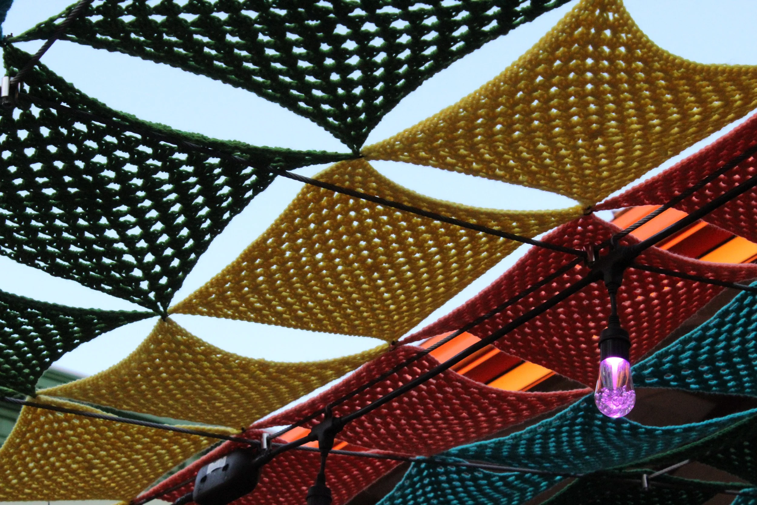 Colorful hanging fabric pieces in green, yellow, red, and blue, arranged in a overlapping pattern with string lights passing through, against a light sky background.