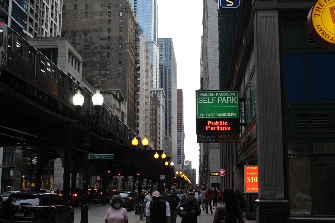 A busy city street with pedestrians, cars, and an elevated train track overhead. There are signs for self parking and public parking, with tall buildings on either side.