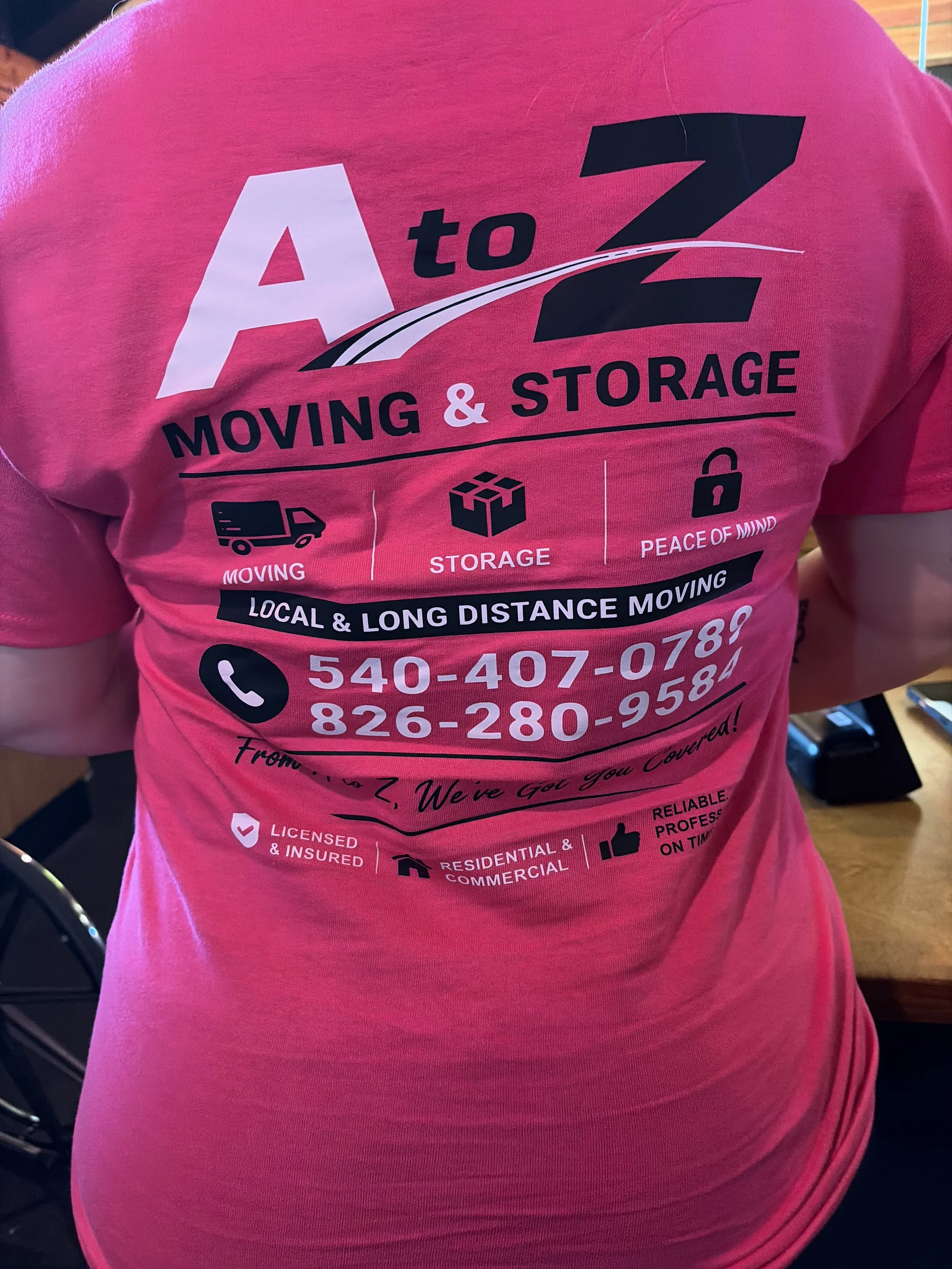 Pink T-shirt advertising moving and storage services with contact information and icons representing moving, storage, peace of mind, and licensing and insurance, worn by a person