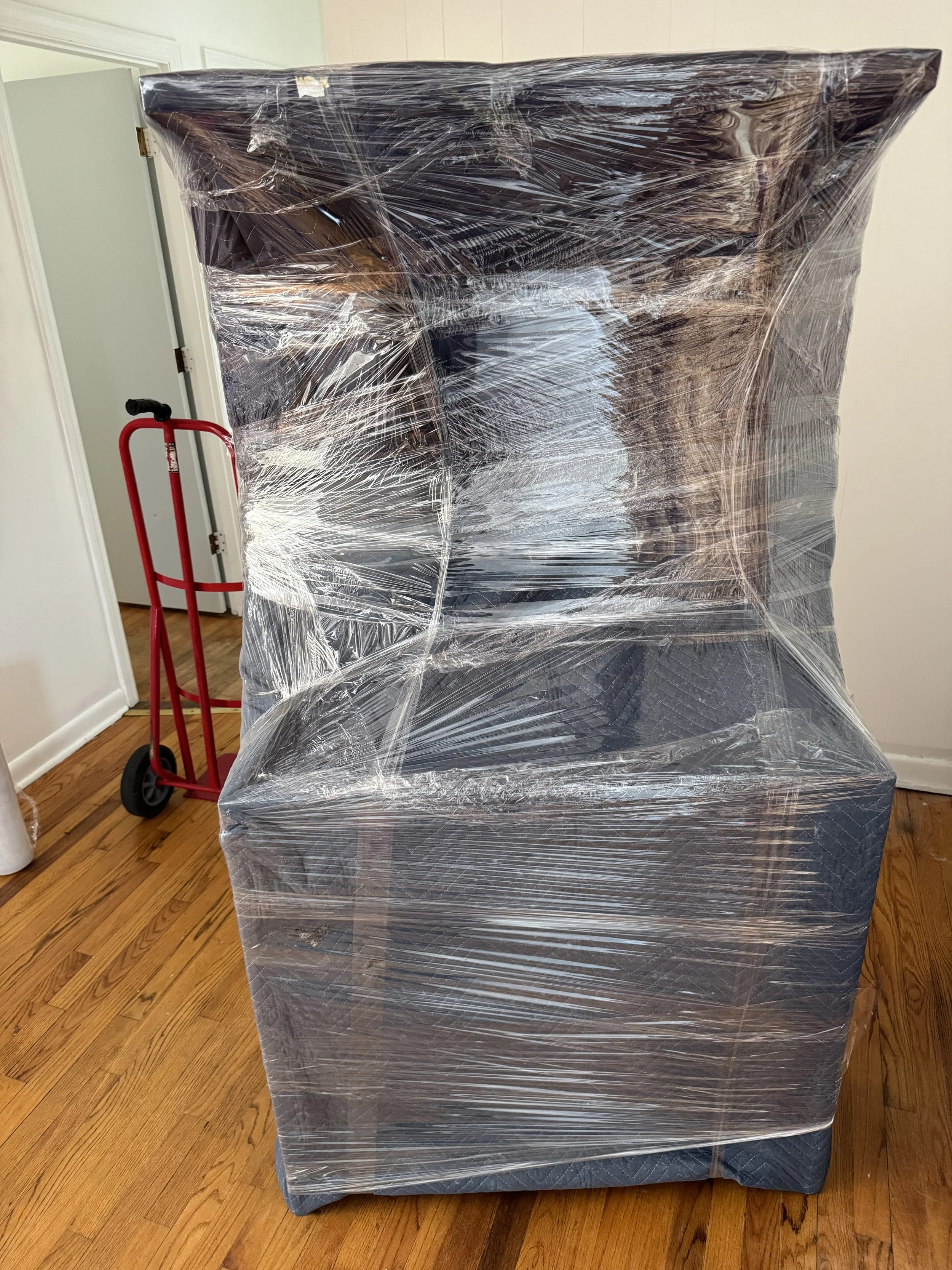 Large piece of furniture, possibly a sofa, wrapped in clear plastic wrap, on hardwood floor inside a room.