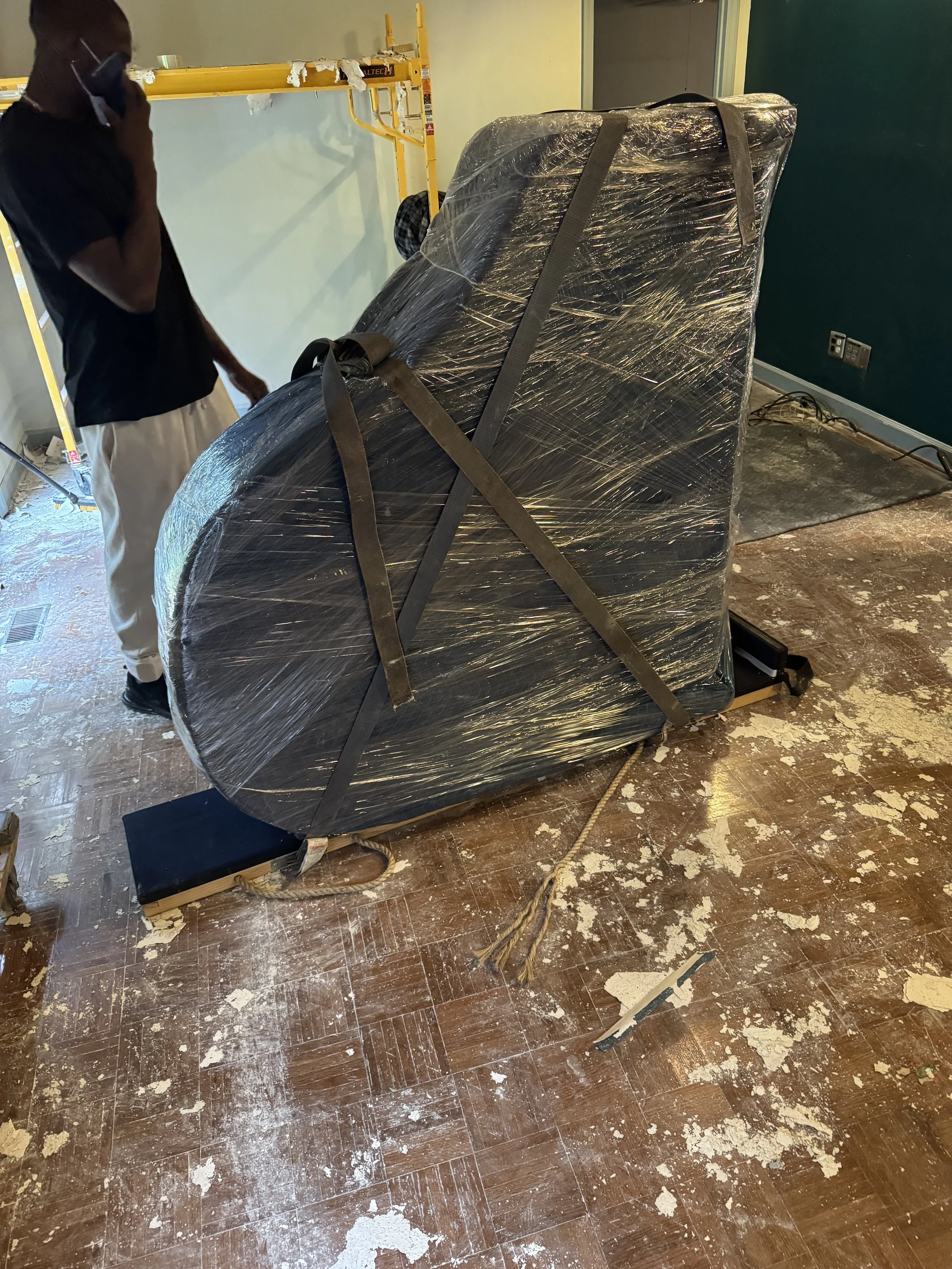 A person on a phone standing beside a large object wrapped in plastic and secured with straps on a wooden floor that is dirty and damaged, with a green wall behind and construction equipment nearby.