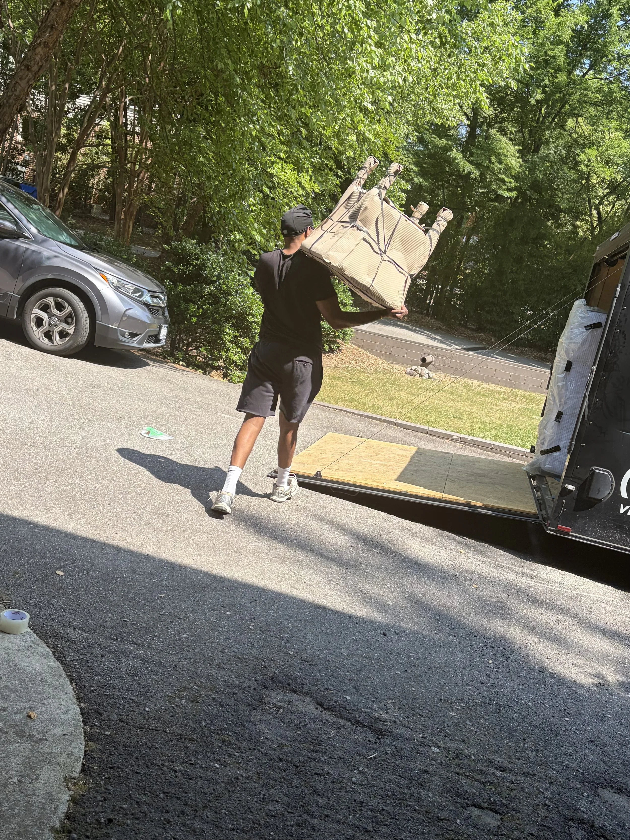 A man wearing a black shirt, shorts, and a cap is carrying a folded outdoor table in a parking lot. There are cars and lush green trees in the background.