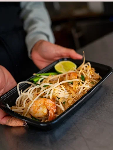 A person holding a black tray of shrimp pad thai garnished with lime and green onion.