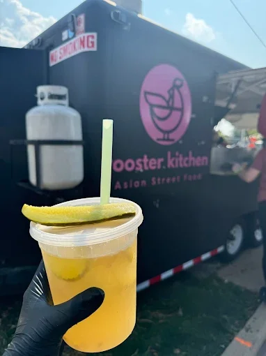 A hand holding a cup of yellow-colored beverage with lemon slices and a straw, in front of a food truck with a logo and sign for Foster Kitchen Asian Street Food.