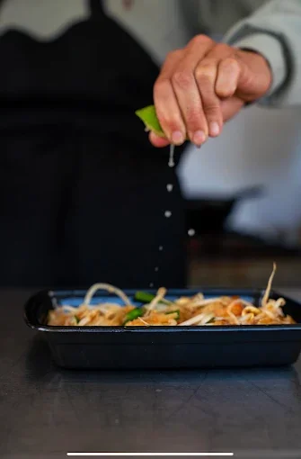 Person sprinkling salt on a dish of noodles with vegetables in a black container.