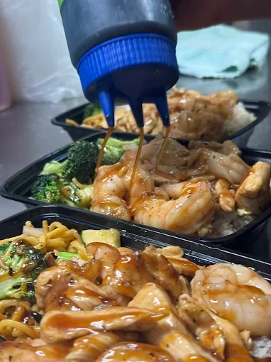 Takeout containers filled with stir-fried shrimp, broccoli, noodles, and other vegetables, with soy sauce being poured on top.