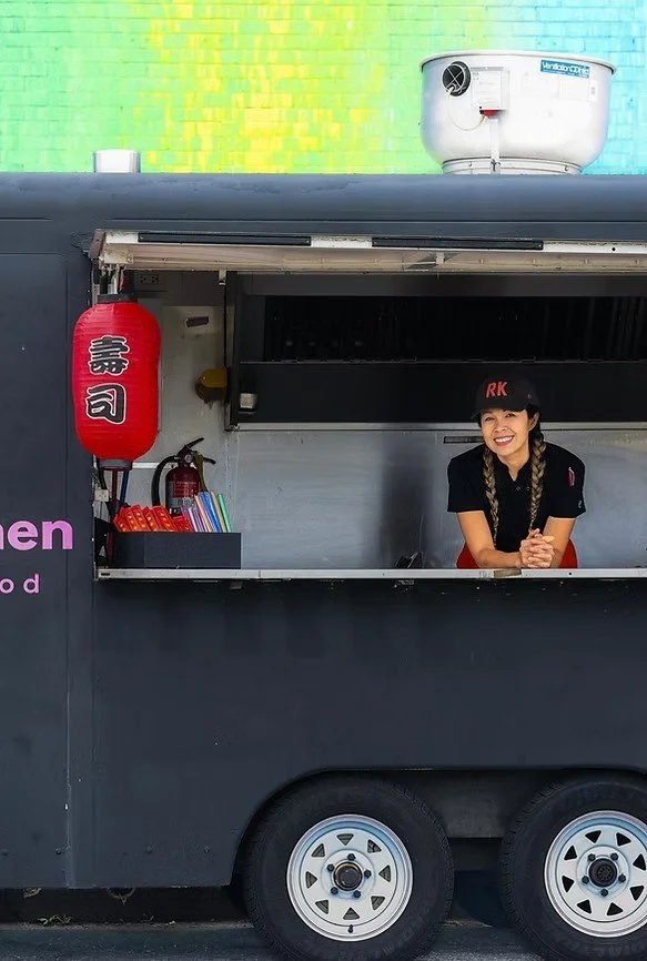 A smiling woman in a black uniform and cap with red letters, standing in a food truck window. The truck has a red Japanese lantern with kanji characters, a small black shelf with colorful items, and a fire extinguisher inside. The background is a colorful wall with a green and yellow pattern.