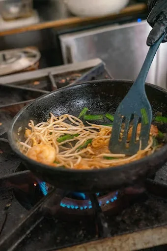 A person cooking noodles and vegetables in a black frying pan over a gas stove.