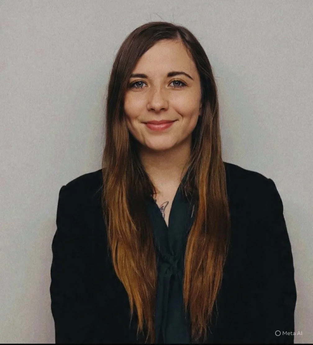 A woman with long brown hair wearing a black blazer and dark blouse, smiling, standing against a plain gray background.