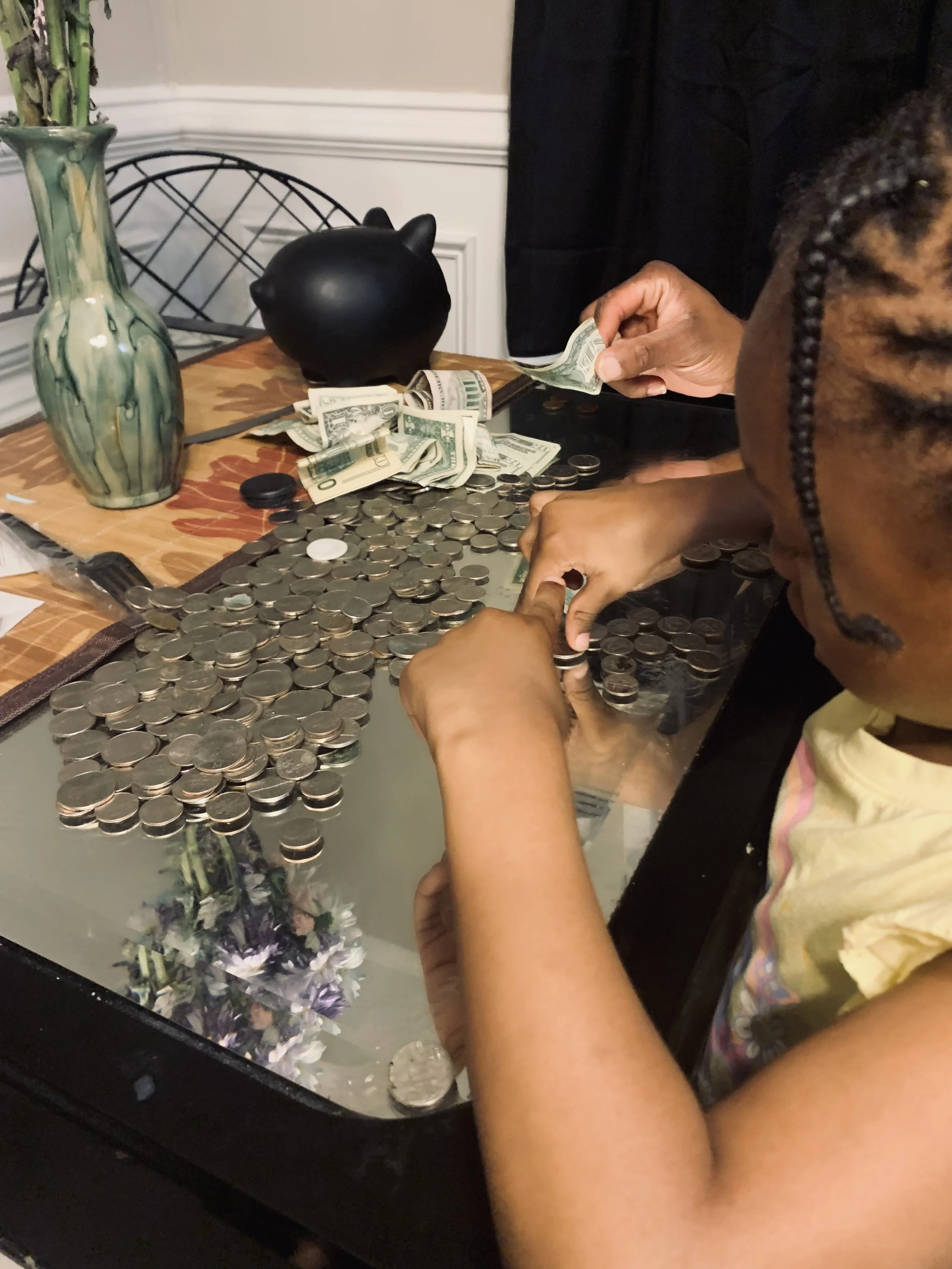 A person counting and organizing cash and coins on a glass table with a flower arrangement, a decorative black piggy bank, and a green vase nearby.