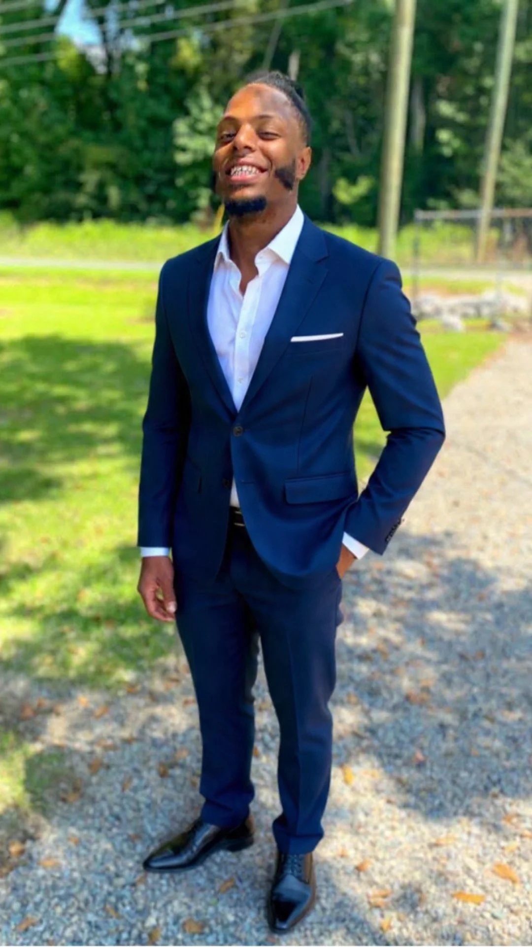 A man in a navy blue suit and white shirt standing outdoors on a gravel path, smiling with one hand in his pocket.