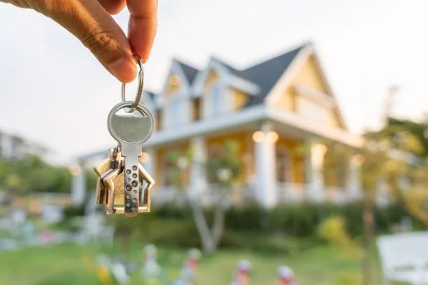 A hand holding a key shaped like a house in front of a large house with a yard.