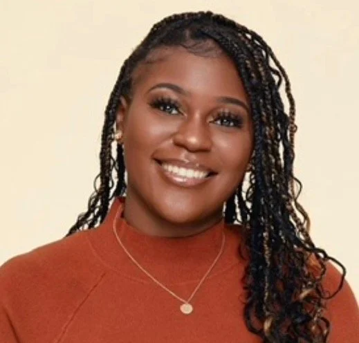 A woman with long, braided hair wearing a rust-colored top and a silver necklace, smiling in front of a plain beige background.