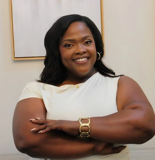 smiling woman with dark hair wearing a white top and gold jewelry crossing her arms in front of a white wall.