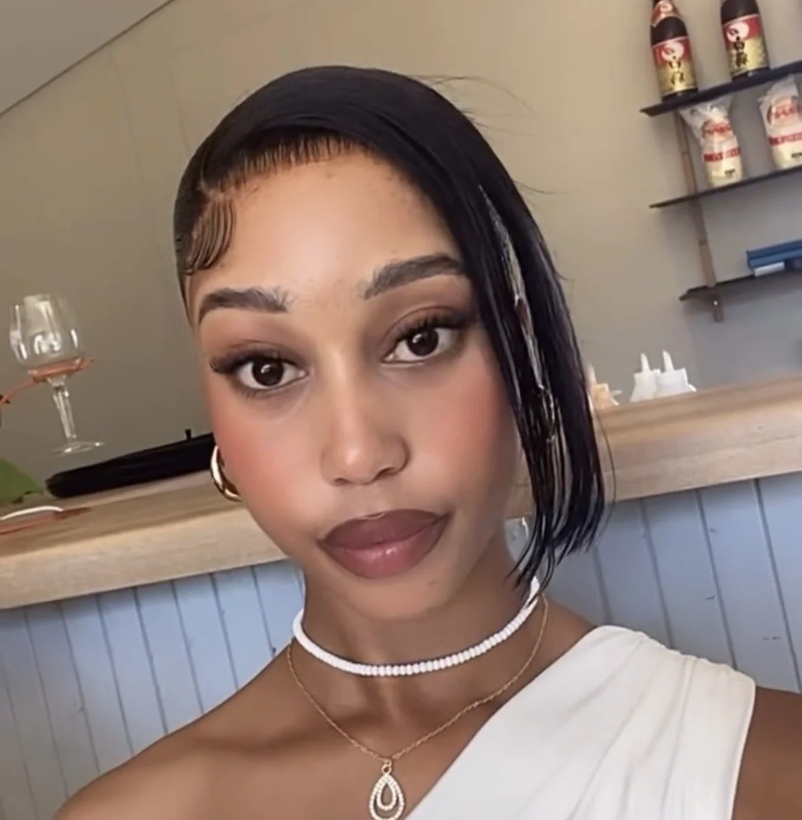A young woman with short black hair, wearing makeup and jewelry, posing indoors with a glass of wine in the background.