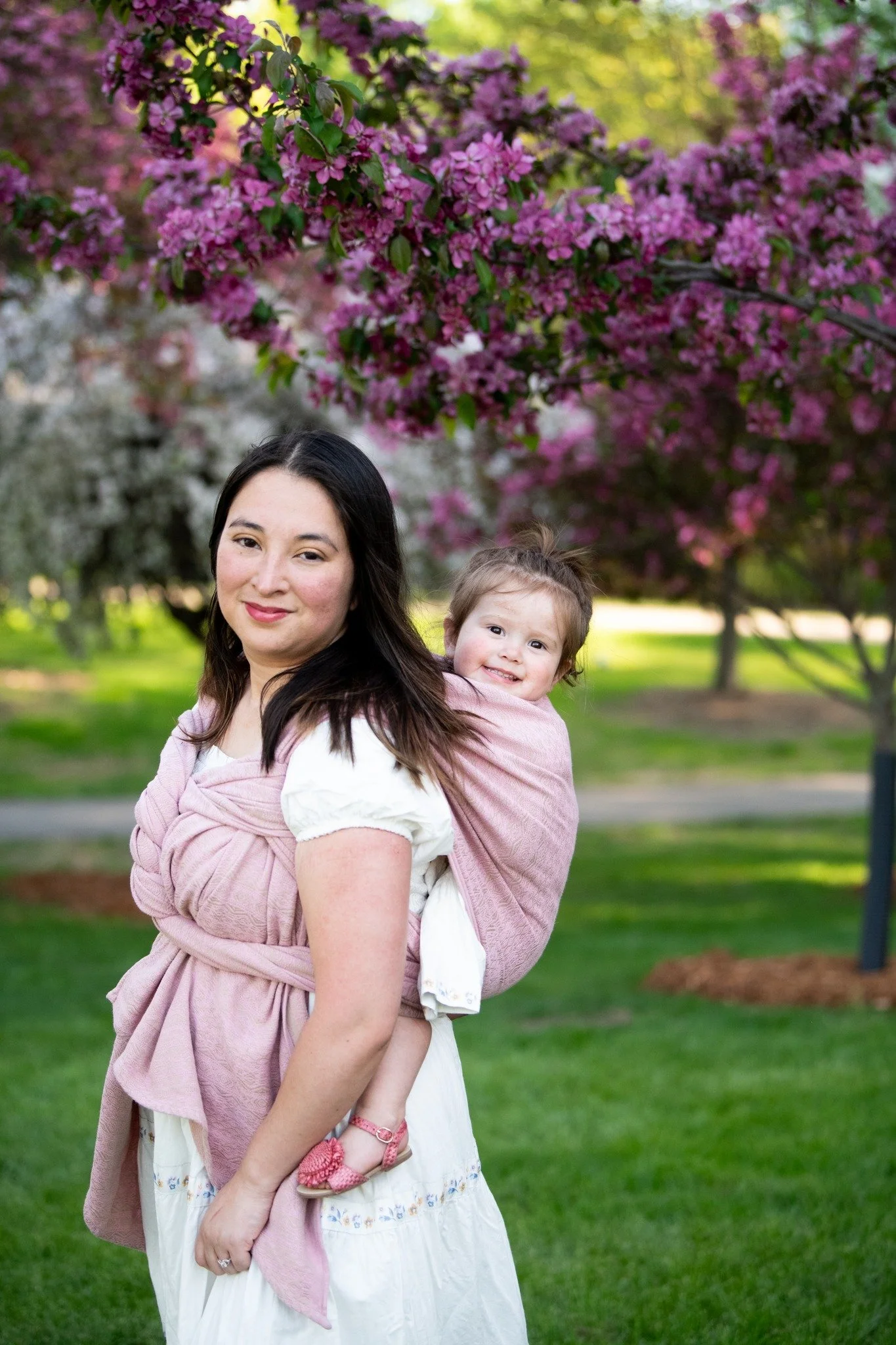 A woman with dark hair holding a young girl with a ponytail, both smiling, standing outdoors with pink flowering trees and green grass in the background.