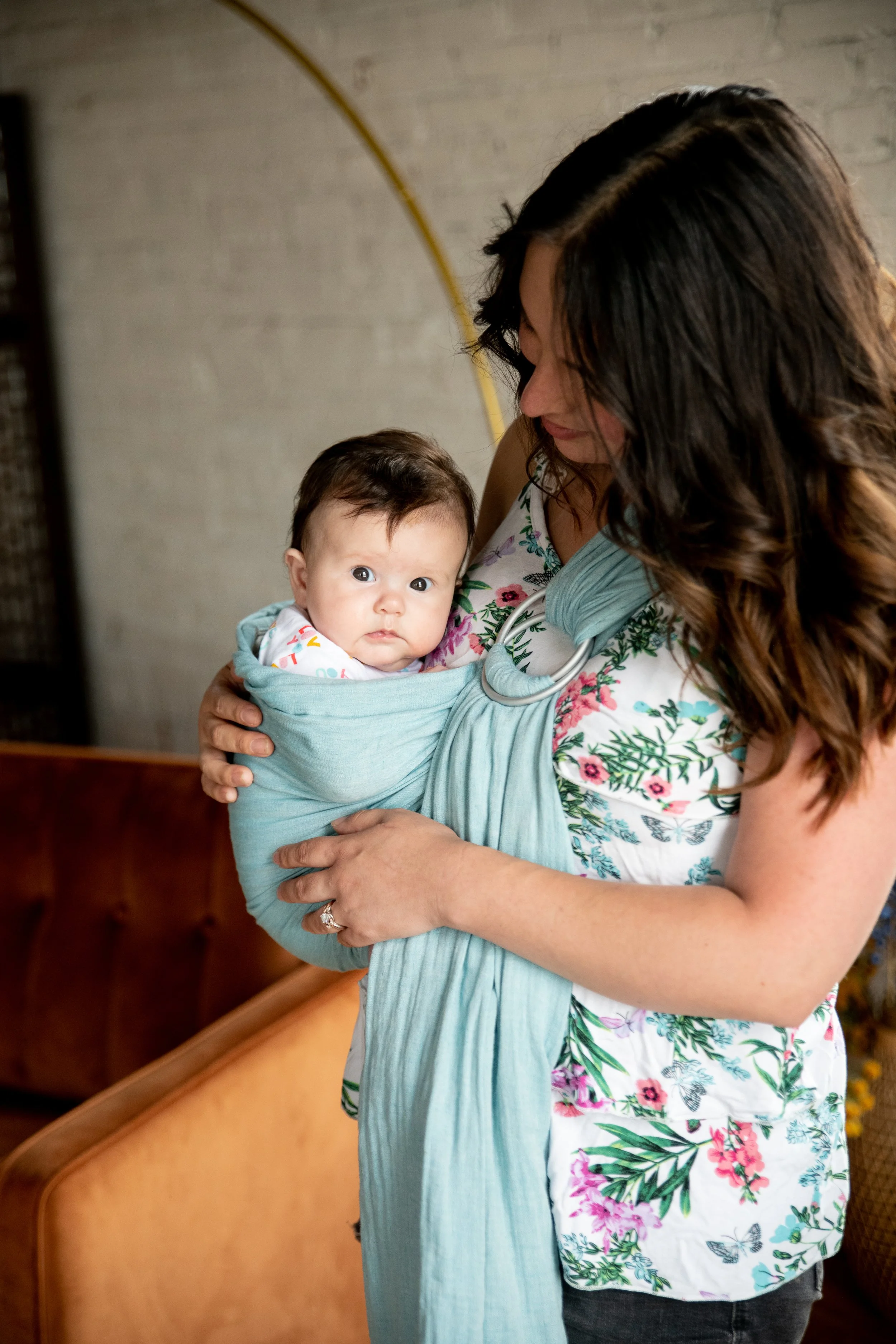 A woman with curly brown hair wearing a floral dress holding a baby wrapped in teal fabric. They are in a room with a beige wall and wooden pews.