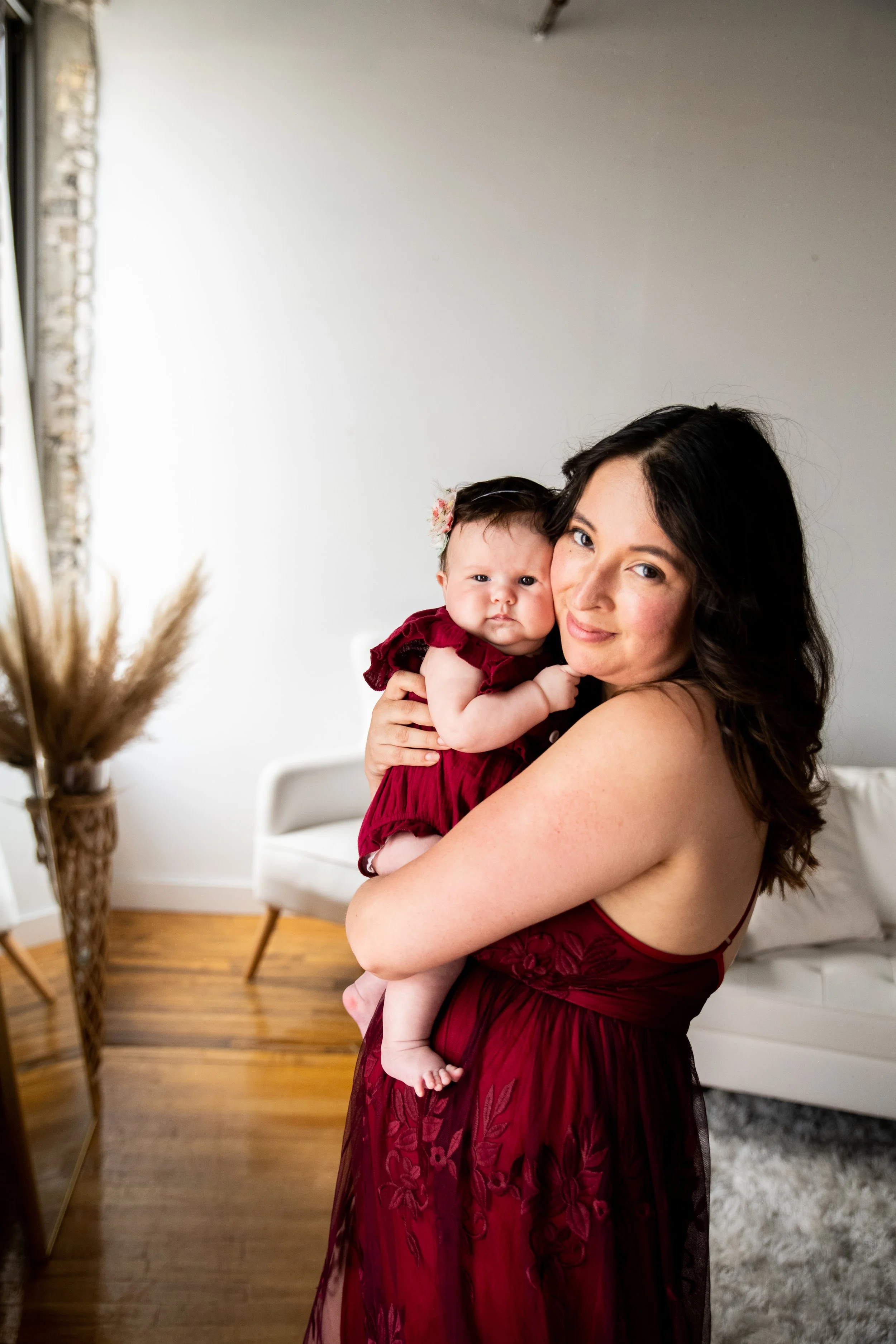 A woman with dark wavy hair and light skin holding a baby girl with dark hair, light skin, and a flower headband, both wearing matching burgundy dresses, inside a home with hardwood floors and a white sofa.