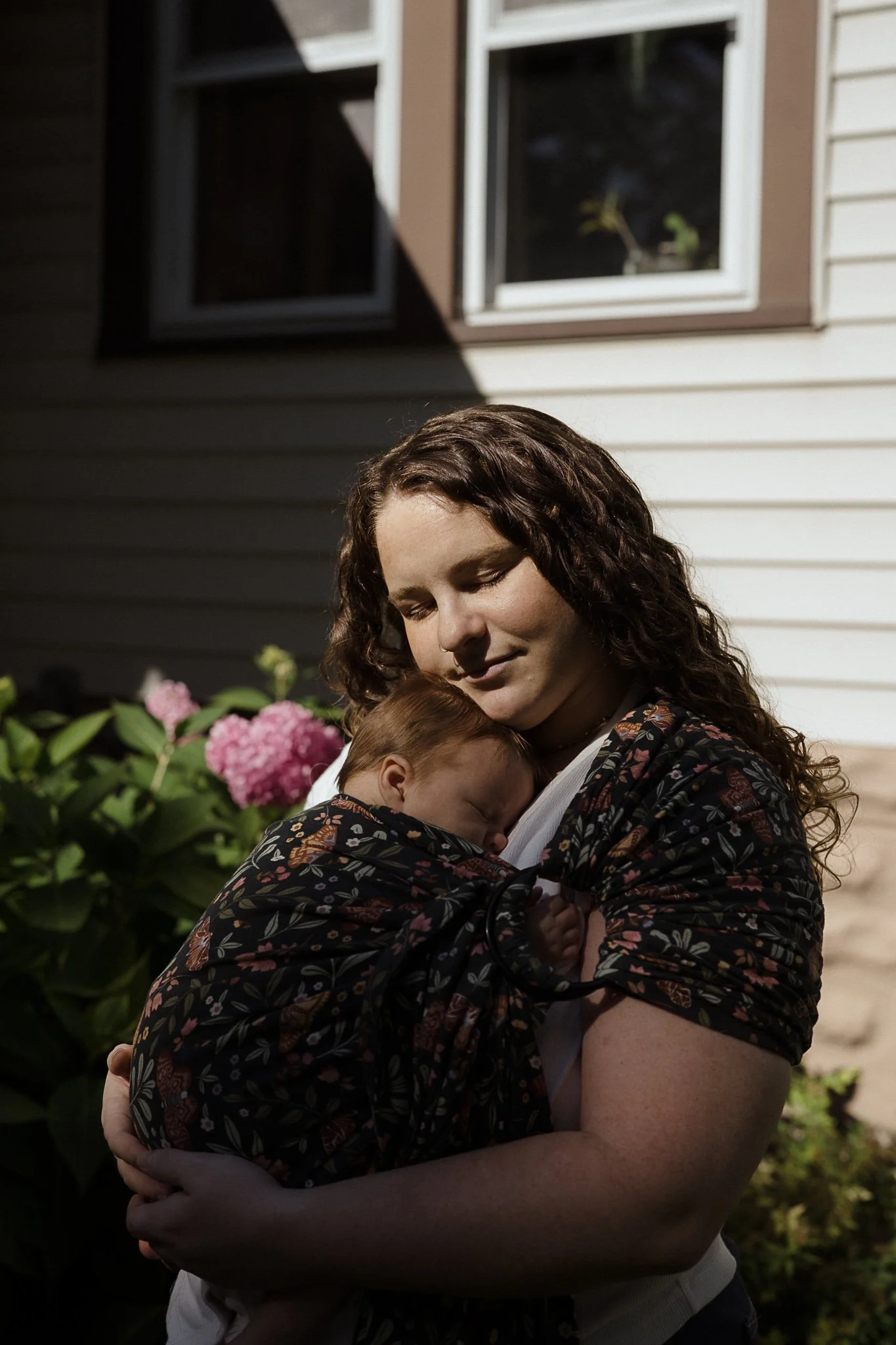 A woman with curly hair holding a sleeping child wrapped in a dark floral ring sling outside near green foliage and pink hydrangea flowers. The background includes a house with white siding and a dark window.