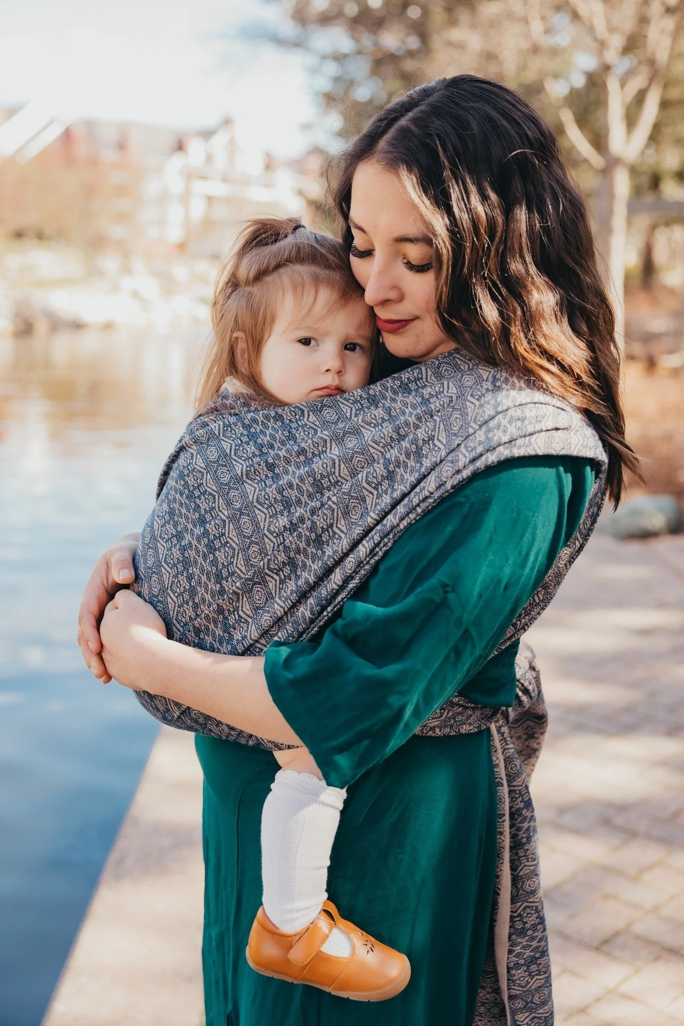 A woman with long dark hair holding a young girl with light brown hair in an outdoor setting near a body of water with trees and buildings in the background.