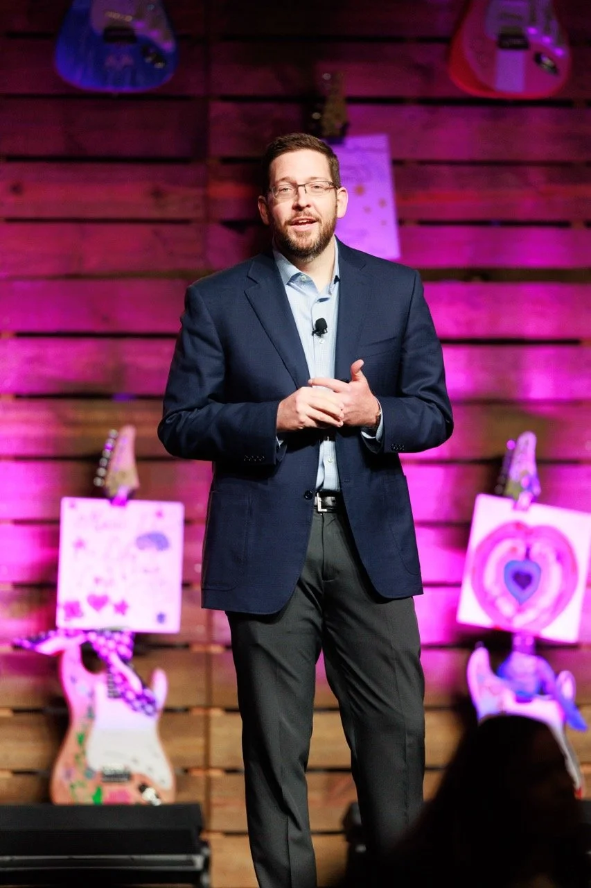 A man in a dark blue blazer and glasses giving a presentation on a stage with pink lighting and decorated with guitars and artwork in the background.
