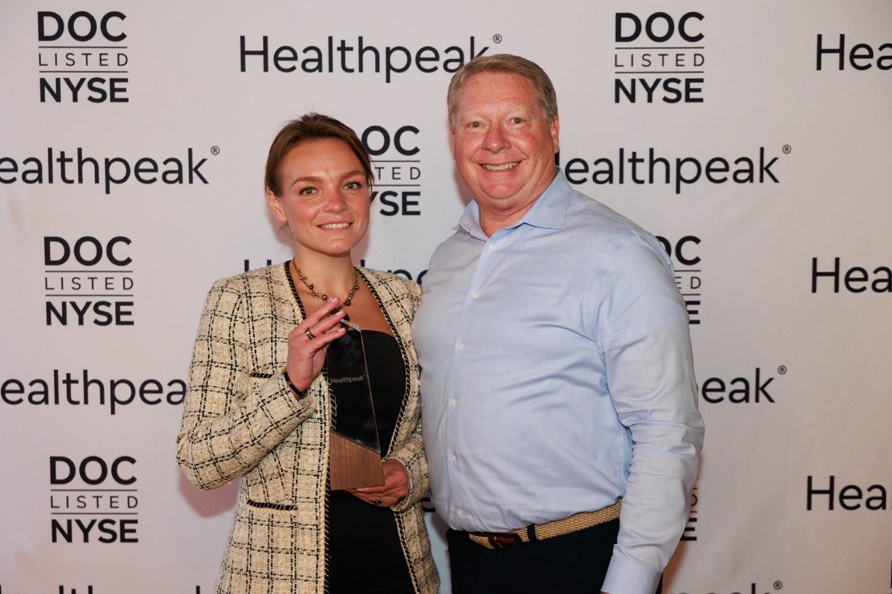 Two people, a young woman and an older man, standing together at a formal event, with a backdrop featuring 'Healthpeak' and 'NYSE' logos. The woman is holding an award and is smiling, while the man is also smiling.