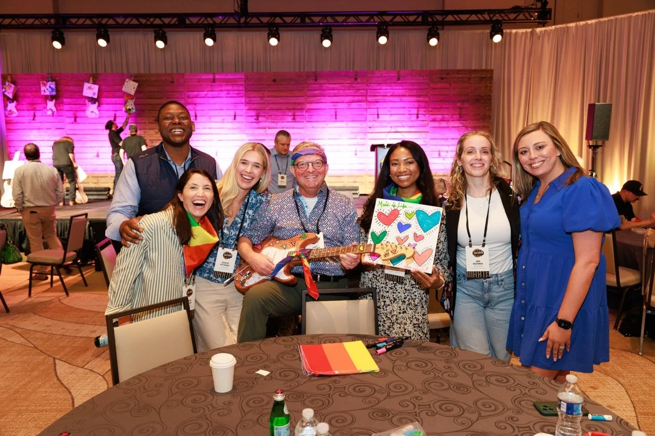 Group of seven smiling people standing around a table at an indoor event, with some holding colorful artwork and a guitar, in a venue with a purple-lit wooden backdrop and stage.