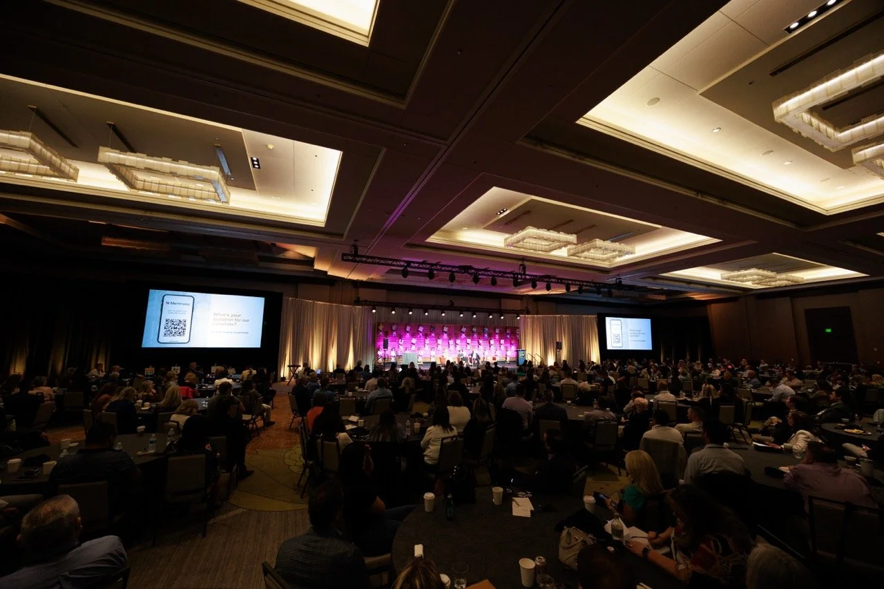 Large conference room filled with seated attendees, presenting on stage with multiple screens, decorative lighting, and a purple backdrop.