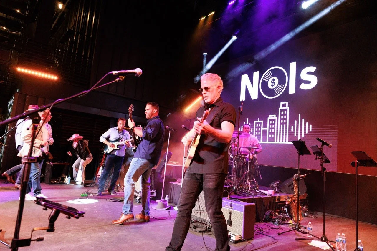 Band performing live on stage with a large screen behind them displaying the word 'NOIS' and cityscape graphics, colorful stage lights illuminating the scene.