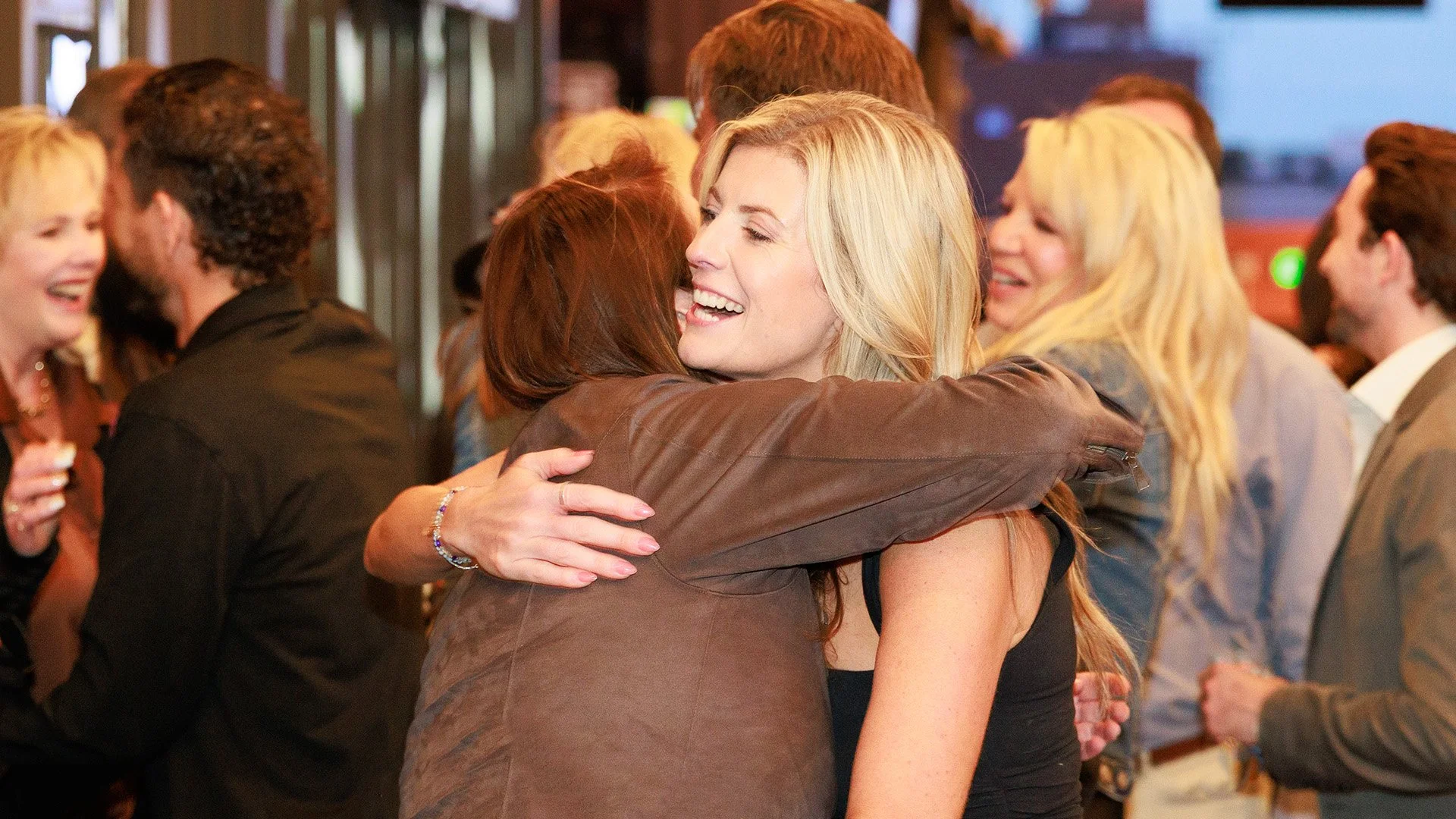 Two women embracing in a hug at a social gathering, smiling happily, with several other people in the background.