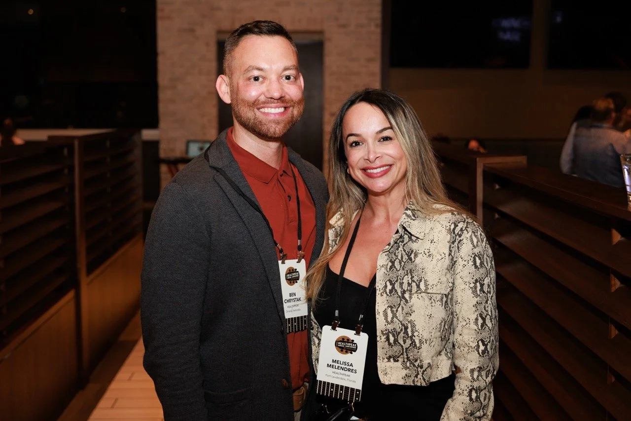 A man and woman smiling at an indoor event, wearing lanyards with name tags, standing side by side.