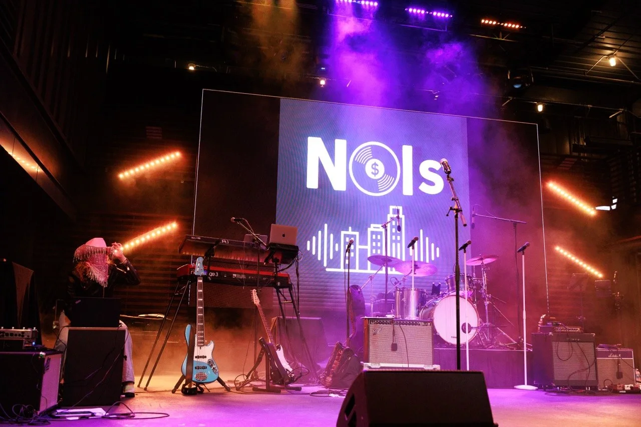 Stage with musical instruments, microphones, a large screen with the word 'NOIS' and a cityscape graphic, purple and orange stage lights, and smoke effects.