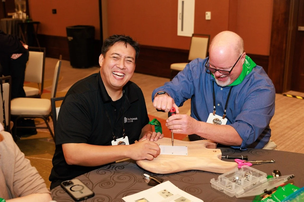 Two men smiling and working together on a woodworking project at a table indoors. One person is using a screwdriver while the other hands him a piece of wood.