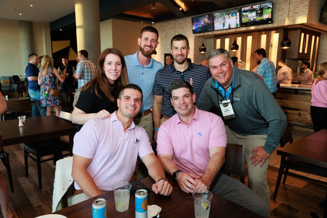 Group of seven people smiling in a restaurant, posing for a photo, with drinks on the table. The background has multiple television screens and other diners.